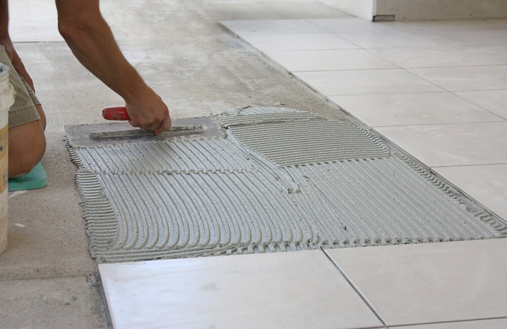 a tiler laying white floor tiles inside of a house using a green knee pad