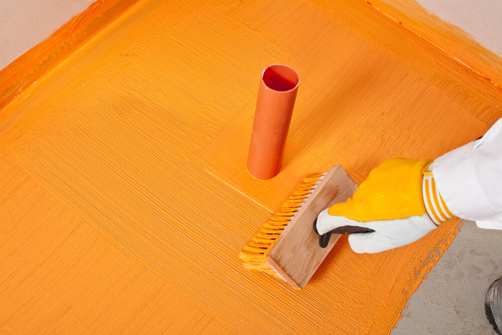 a tiler painting on orange waterproof paint around a pipe that's coming out of the ground