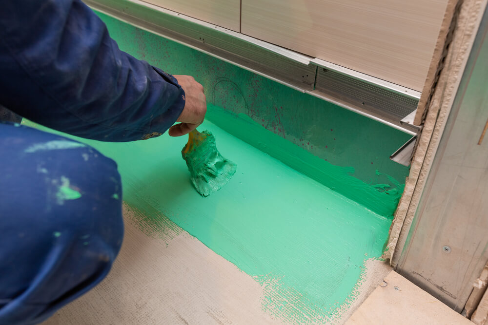 a tiler painting on some green waterproof paint at the bottom corner of a bathroom, under some cupboards 